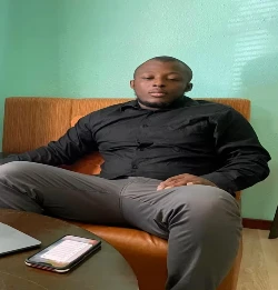 Jhon Arzu-Gil, an Application Developer at IBM, sitting in a chair, dressed in a black shirt and gray pants, with a smartphone and laptop on a table beside him.